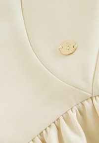 Kremfarvet stof med rynket detalje og en lille oval guldfarvet charm med bogstavet B indgraveret, syet fast på materialet.