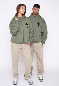 Felpe con cappuccio zip olive verdi con ricamo a croce nero, abbinate a pantaloni della tuta beige. Entrambi i capi presentano una vestibilità comoda e un tessuto morbido.
