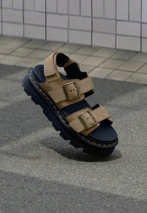 Sandale en daim beige avec semelle épaisse noire, brides ajustables avec boucles dorées, flottant au-dessus d'un trottoir gris près d'un mur carrelé.