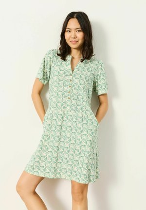 Žena s tmavými vlasmi po plecia nosí zelené vzorované šaty s krátkym rukávom na gombíky a s vreckami, stojaca pred jednoduchým pozadím.