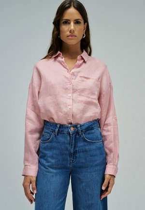 Mujer con cabello largo y castaño, vestida con una camisa rosa claro abotonada metida en unos vaqueros azules de talle alto, sobre un fondo liso.