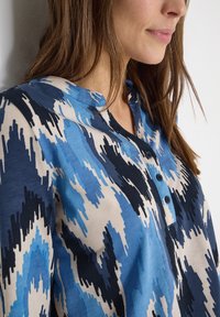 Femme portant un chemisier à motifs bleu, noir et beige avec un col rond et trois boutons bleu marine, visible des épaules au menton.