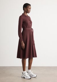 Rochie bordeaux, cu maneci lungi, guler și nasturi, talie strânsă și fustă evazată; asociată cu ciorapi albi și adidași albi sport.