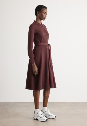 Bordeaux kleurige jurk met lange mouwen, kraag en knopen, een getailleerde taille en een uitlopende rok; gecombineerd met witte sokken en sportieve witte Sneakers.