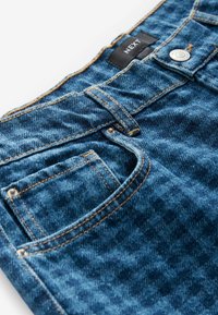 Gros plan sur un jean en denim bleu montrant la poche avant, la lanière de ceinture, les rivets en métal, le bouton et l'étiquette en tissu noir avec le texte "NEXT".