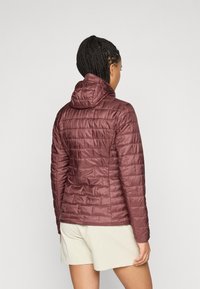 Femme avec des cheveux tressés portant une veste à capuche matelassée bordeaux et un short beige clair, se tenant devant un fond blanc uni.