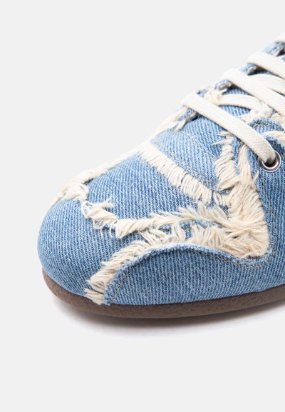 Gros plan sur l'avant d'une chaussure en denim bleu avec un motif en tissu beige effiloché et des lacets blancs sur un fond blanc.
