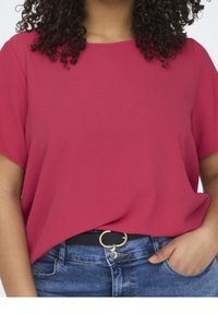 Woman with curly hair wearing a short-sleeve pink top tucked into blue jeans with a black belt, hand in pocket.