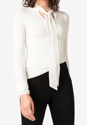 Blusa bianca a maniche lunghe con un nodo al collo e dettagli cut-out. Realizzata in tessuto morbido, di stile aderente, abbinata a pantaloni neri.