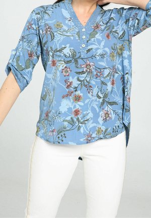 Femme portant un chemisier bleu à fleurs boutonné avec manches retroussées et pantalon blanc, posant avec un bras levé sur un fond uni.