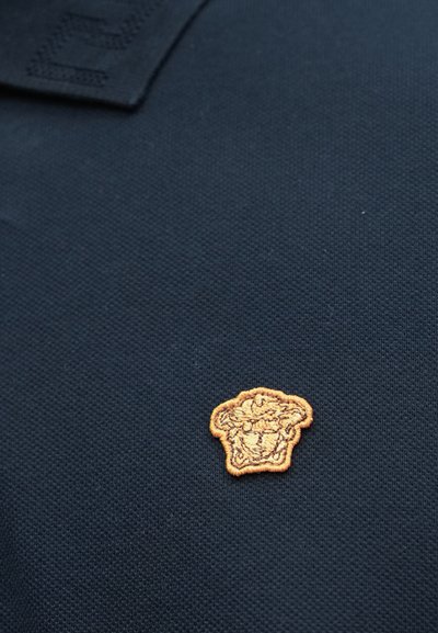 Πόλο μπλούζα σε ναυτικό μπλε με υφή υφάσματος, διακοσμημένη με κεντητό χρυσό λιοντάρι στο αριστερό στήθος.