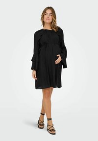 Vestido de maternidad negro con mangas acampanadas y cintura atada. Tejido ligero con forma fluida, combinado con sandalias negras de tiras.