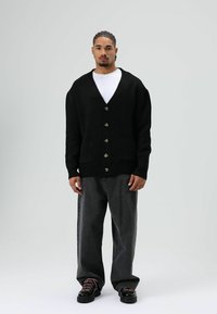 Cardigan en maille noire avec col en V, six boutons et deux poches avant. Porté sur un T-shirt blanc, associé à un jean gris ample et des chaussures noires.