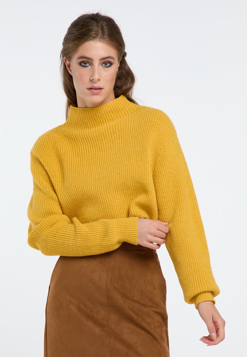 Geel geribde trui met een hoge kraag en losse mouwen, gecombineerd met een bruine leren rok, wat een textuurcontrast creëert.