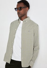 Camicia a quadri verde e bianca con colletto button-down sopra una maglietta bianca. Presenta una piccola tasca ed è indossata con occhiali da sole scuri.