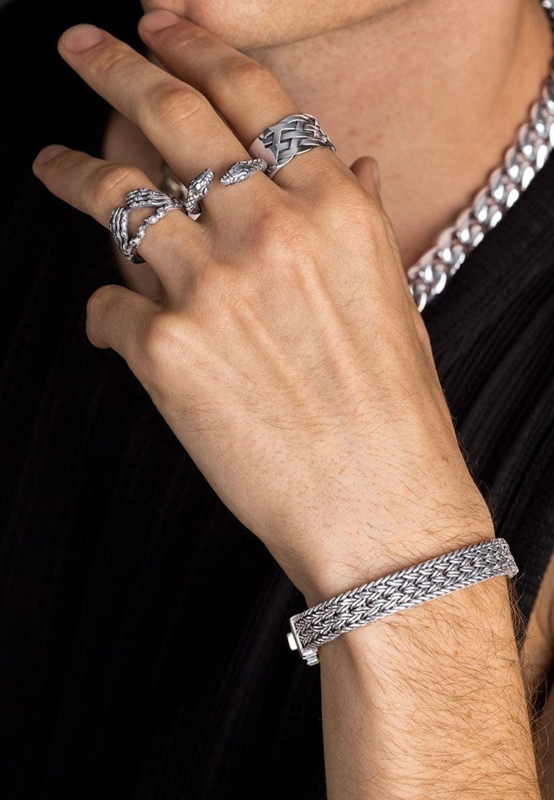 Silbernes Schmuckstück mit mehreren Ringen und einem geflochtenen Handgelenkband. Die Ringe zeigen komplexe Designs, während das Armband eine strukturierte Oberfläche hat.