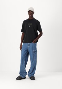 Svart bomull T-skjorte med "GUESS" logo, lyseblå denim cargo bukser med sidelommer, og svarte sneakers. Tilbehør: beige cap.