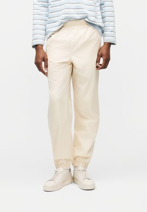 Ember krémszínű gumis derekú melegítőnadrágot, krémszínű tornacipőt és világoskék-fehér csíkos hosszú ujjú inget visel, és egyszínű háttér előtt áll.