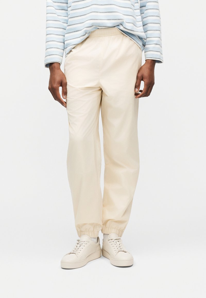 Άτομο που φοράει κρεμ παντελόνι jogger με ελαστική μέση, κρεμ αθλητικά παπούτσια και ένα μακρυμάνικο πουκάμισο με γαλάζιες και λευκές ρίγες, στέκεται μπροστά σε απλό φόντο.