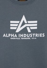 Sivi dukserica s velikim bijelim grafičkim logotipom sa stiliziranim slovom "A" i tri horizontalne pruge, natpis "ALPHA INDUSTRIES."