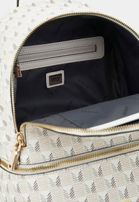 Sac à dos avec un motif géométrique en beige et marron. Matériau lisse, fermeture éclair dorée, deux poches intérieures, étiquette de marque et doublure grise.