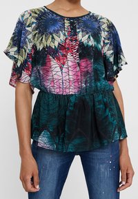 Blusa con fantasia floreale e maniche corte a volant. I colori includono blu, rosa, verde e nero. Presenta un punto vita arricciato e un tessuto leggero.