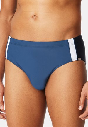 Mann trägt blaue Badehose mit weißen Seitenstreifen, gezeigt von der Taille bis zur Mitte des Oberschenkels vor einfarbigem Hintergrund.