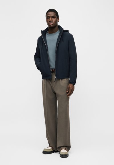Νεαρός άνδρας όρθιος, φοράει μπλε ναυτικό μπουφάν με φερμουάρ, μπλε-γκρι φούτερ, καφέ φαρδιά παντελόνια και μπεζ loafers με μαύρες σόλες.