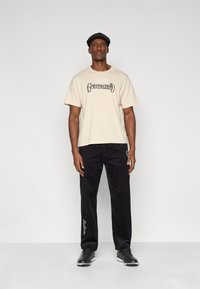 T-shirt en coton beige avec texte noir "Grateful Dead", associé à un pantalon en velours côtelé noir avec broderie blanche et coupe droite.