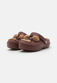 Zoccoli Croslite marroni con superficie texturizzata, dotati di due pompon pelosi e di una decorazione nera sul davanti. Design aperto sul retro.