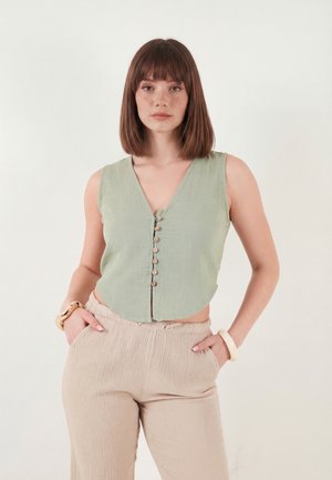 Noor naine sileda pruunide juustega, kes kannab varrukateta salvei rohelist nööbitavat toppi ja beeže tekstuuriga pükse, käed taskutes, neutraalne taust.