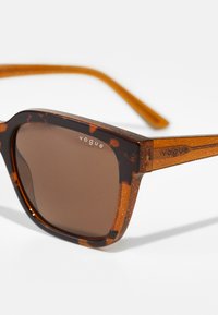 Schildpattsonnenbrille mit braunen Gläsern, glitzernder Textur, quadratischer Form und silbernem "Vogue"-Logo auf dem Gestell.