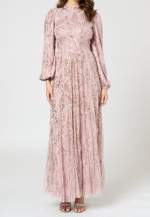 Femme portant une longue robe rose à manches longues avec des motifs en sequins argentés, associée à des sandales argentées ouvertes à l'avant.