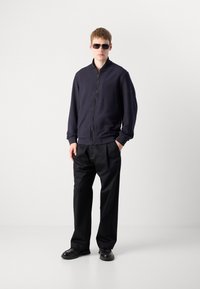 Jongeman met donkere zonnebril, een marineblauwe zip-up jas, zwarte rechte broek en zwarte schoenen, staande tegen een effen witte achtergrond.