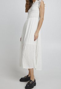 Biała midi sukienka z marszczonym nadrzędzeniem, falbaniastymi rękawami i teksturowanym materiałem z małymi wypukłymi kropkami; zestawiona z czarnymi platformowymi mokasynami.