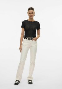 Svart teksturert kortermet topp med bølgete kanter, paret med hvite vide jeans og et svart belte med sirkulære sølvdetaljer.