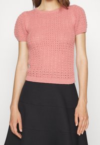 Top de punto rosa de manga corta con patrón texturizado y detalles de canalé. Presenta una cintura ajustada y mangas abullonadas, combinado con una falda negra.