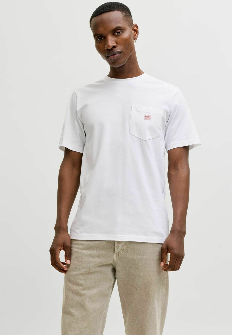 Mand iført hvid t-shirt med korte ærmer og brystlomme samt lyse beige bukser, stående mod en ensartet lys grå baggrund.