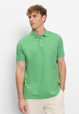 Man die een groene polo met korte mouwen en lichtbeige broek draagt, staat met één hand in de zak en kijkt naar links.