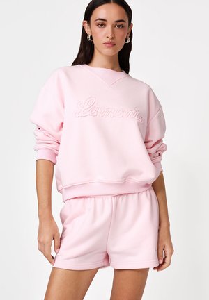 Kobieta w jasnoróżowej bluzie z napisem "La Martina" i pasujących szortach, stojąca na jednolitym jasnym tle.