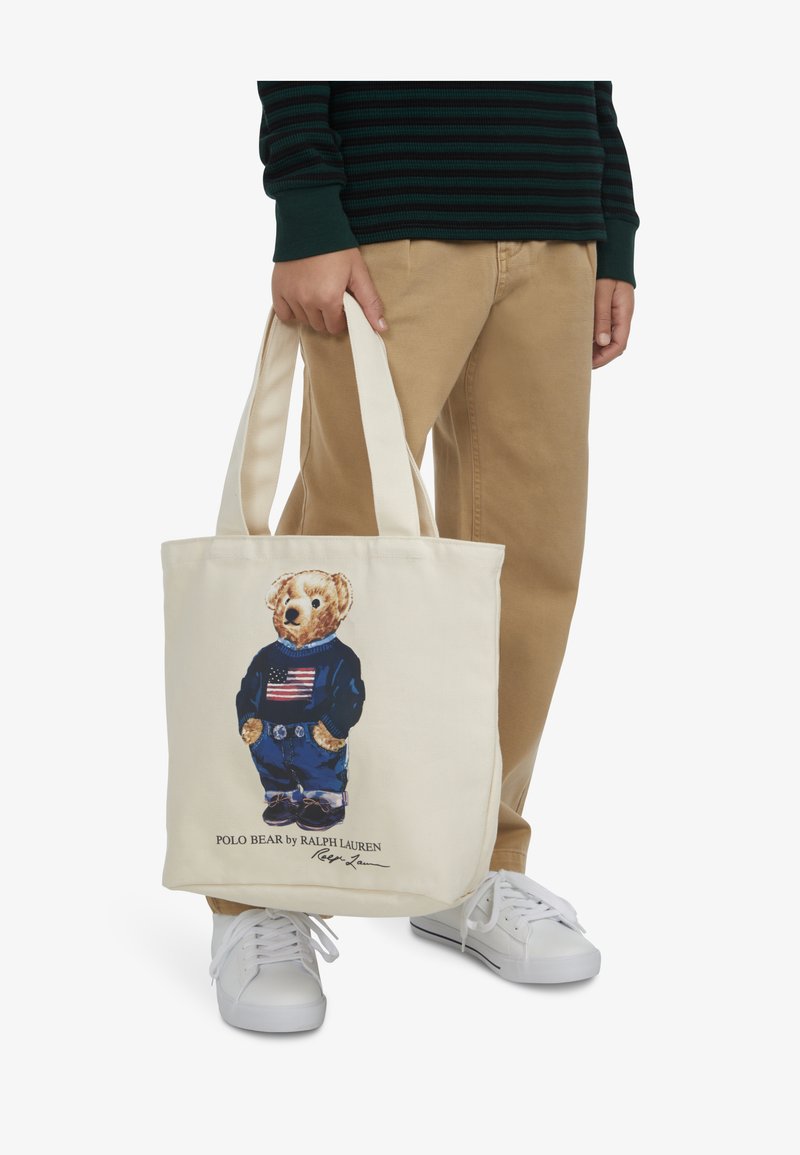 Kanvasova torba v kremni barvi z grafiko medveda v mornarsko modrem puloverju z ameriško zastavo, modrimi kavbojkami in teniskami.