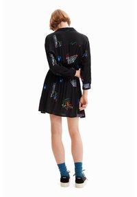 Abito nero con maniche lunghe, stampe grafiche colorate, colletto e orlo svasato. Combinato con calzini blu e scarpe nere.