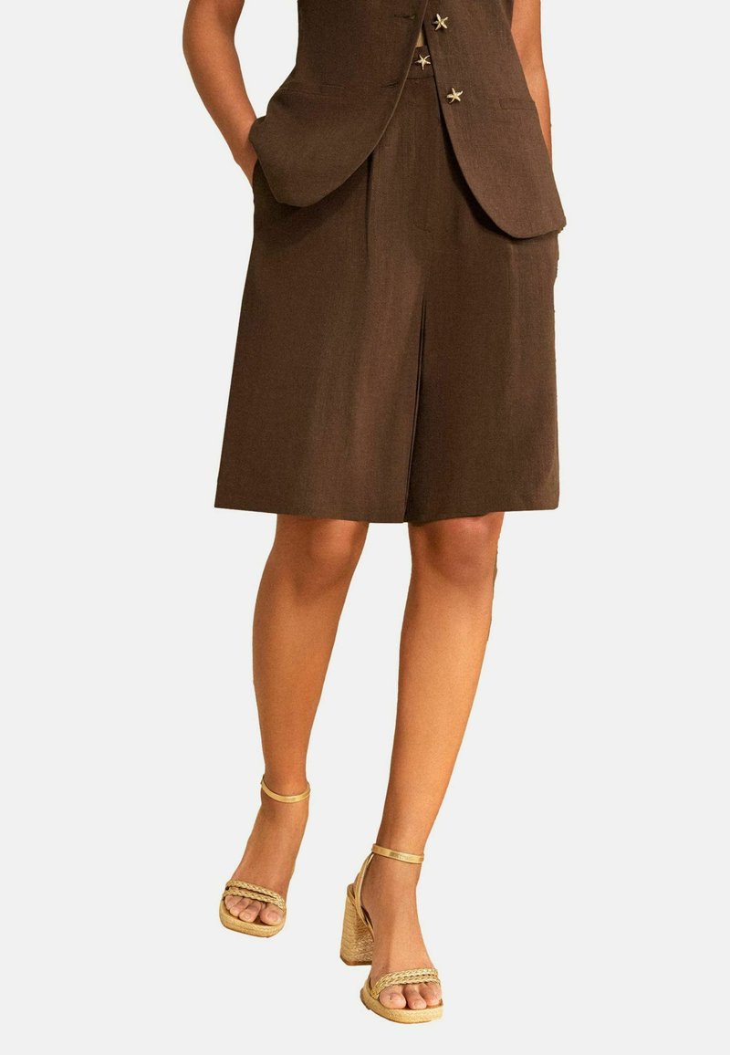 Brauner knielanger Rock mit Frontschlitz, passende Weste mit sternförmigen Knöpfen, Hände in den Taschen und beige Sandalen mit Absatz.