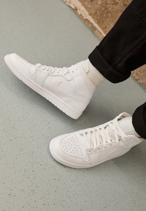 Person iført hvide høje sneakers med snørebånd og hvide sokker på en lys grå, plettet gulv, iført sorte bukser.