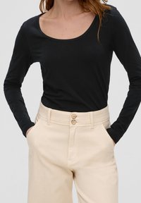 Schwarzes Langarmoberteil mit weitem Rundhalsausschnitt, kombiniert mit beigen, hoch taillierten Hosen mit zwei Knopfverschlüssen und Seitentaschen.