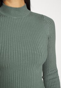 Langsleeve, geribbelde coltrui in een gedempt groene kleur. Kenmerken verticale strepen en een strakke pasvorm met een hoge kraag. Zachte textuur.