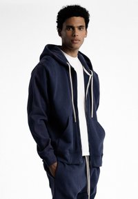 Sweat à capuche zippé bleu marine en tissu mélangé coton. Doté de poches avant, d'une capuche avec cordon de serrage et de poignets côtelés. Porté avec une chemise blanche.