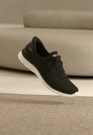 Zapatilla de punto negra con suela blanca, cordones y logotipo de Skechers en la lengüeta, flotando sobre una alfombra beige frente a un sofá beige.