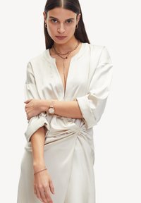 Silke bluse i creme med V-udskæring, løse ærmer og en twist-detalje ved taljen. Accessories inkluderer et lagdelt kædehalssmykke og armbåndsur.