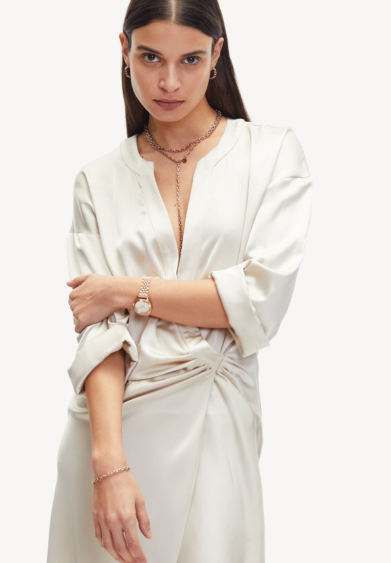 Silke bluse i creme med V-udskæring, løse ærmer og en twist-detalje ved taljen. Accessories inkluderer et lagdelt kædehalssmykke og armbåndsur.